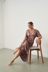 Elegant woman in a mauve dress sits gracefully on a wooden chair, her legs crossed. She wears...