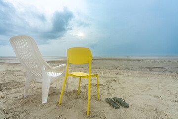 Empty white yellow plastic chairs on deserted sandy beach under cloudy sky, with flip-flops. Evokes...