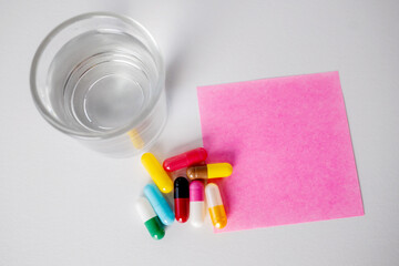 Top view of glass of water, many pill capsules and pink note paper on white background with copy space.