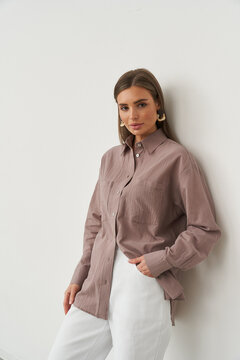 Woman leaning against wall wearing long-sleeved, button-up, mauve shirt and white pants, gazing at camera with calm expression. Her hair is styled straight, accessorized with round earrings
