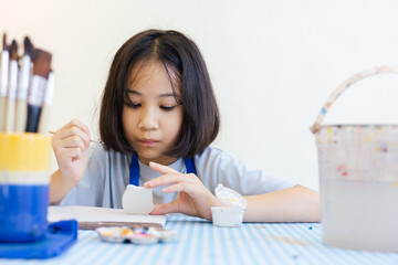 Young Asian girl focuses on painting a clay object, with her hand and brush with selective focus shows creativity and calm concentration for kid learning activity  via art and development in child.