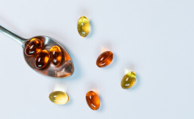 A close-up, top-view image of golden fish oil capsules on a silver spoon, isolated on a clean white background. This flat lay composition represents the concept of daily nutritional supplements.