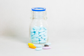 Glass bottle filled with blue pills, with colorful tablets scattered around on a clean white background, representing modern pharmaceutical concept and daily supplement usage in healthcare.