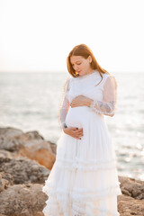Smiling pregnant woman 25-29 year old in vintage white dress holding tummy standing over sea and...