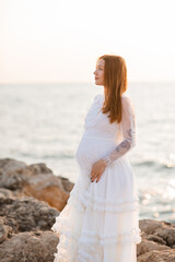 Beautiful pregnant woman 25-29 year old in vintage white dress standing over sea and sunset outdoors. Maternity wear. Summer vacation season