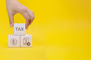 A hand stacks white blocks with the word TAX and financial icons against a yellow background, representing tax planning.