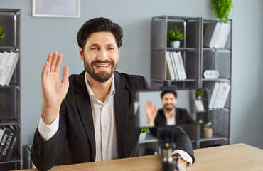 Cheerful man dressed in formal suit, shooting video blog on camera while posig at workplace, waving...