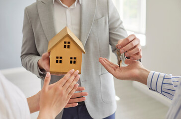 Closeup of realtor hands holding small wooden house and giving key to a couple customers standing...