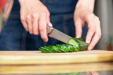 Close-up of knife cutting juicy bright cucumber on wooden cutting board. Preparing ingredients for cooking. Healthy and wholesome food concept
