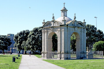 Large historic stone gate with arches and dome, surrounded by trees and green grass on a sunny day....
