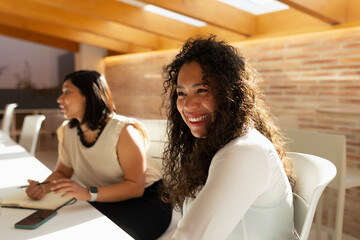 Two young women, dressed in a casual but formal way, and of Latin and Asian features, laugh during an office meeting