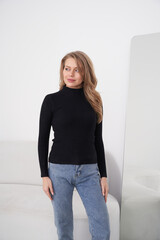 A woman with long, wavy hair wears a black ribbed turtleneck and blue jeans, standing near a mirror...