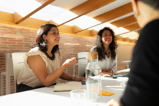 Two young women, dressed in a casual but formal way, and of Latin and Asian features, laugh during an office meeting