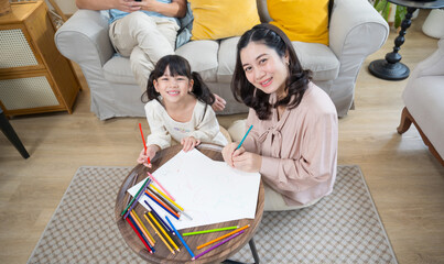 Asian mother and daughter happily sit together in the living room, drawing pictures and enjoying...