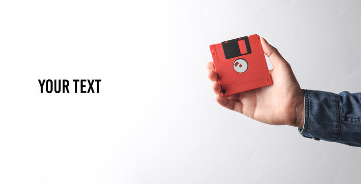 Man's hand in denim shirt holding red floppy disk on gray background. Copy space for your text