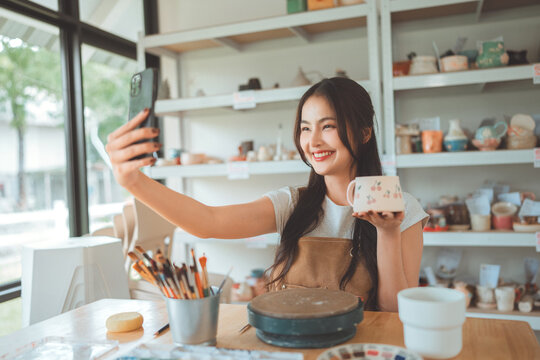 Happy asian female artist taking selfie with handmade pottery design in creative studio – smiling sculptor sharing artwork on social media for online selling, digital portfolio and creative expression