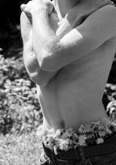 Close-up of a shirtless person hugging themselves, with flowers tucked into jeans