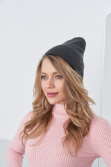 Young woman with long blonde hair wears a dark gray beanie and light pink ribbed top, looking to...