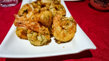 Close-up of golden, crispy, deep-fried shrimp coated in a light, herb-infused batter, arranged on a white rectangular dish over a red tablecloth