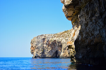 Fototapeta premium ZURRIEQ, MALTA - Augusts 06, 2021: The Blue Grotto - A famous sea cave surrounded by the deep blue sea at southern Malta. On a traditional boat surrounded by more brightly coloured boats. On a hot day