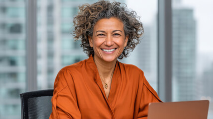 Smiling mature woman with curly hair wearing orange blouse working on laptop in office