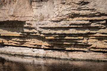 The yellow limestone wall of a canyon is cut by a stream flowing at its base. Sharp shadows...