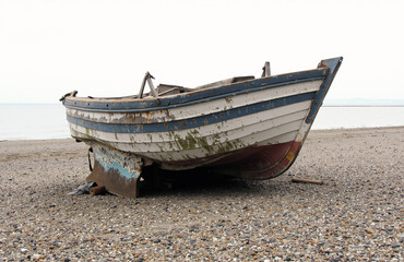 Naklejka premium Abandoned fishing boat on pebbly beach under overcast sky.