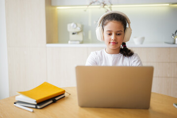 Engaging young girl with headphones immersed in online learning while seated at a wooden table in a...