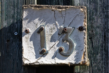 Weathered metal house number 13 mounted on a cracked, aged concrete plaque, attached to a rustic...