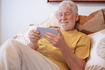 Happy senior white-haired man sitting on bed using smartphone enjoying tech and social. Elderly smiling grandfather using phone