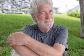 Portrait of attractive smiling relaxed senior man of 70 years old sitting in the meadow looking away. Elderly grandfather with white hair and beard enjoying retirement lifestyle