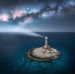 Aerial view of lighthouse on smal island in the sea and Milky Way at night in summer. Beautiful lighthouse with light beam, blue sky with stars. Landscape. Adriatic sea, Croatia. Space. Top view