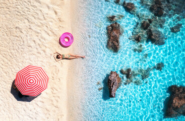 Aerial view of the lying beautiful woman with pink swim ring on the sandy beach, blue sea, stones,...