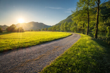 Sun setting behind mountains illuminating gravel road winding through green grass field next to a...