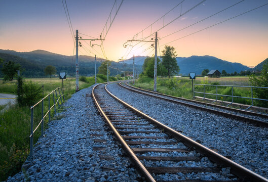 Empty railroad tracks disappearing into the sunset in a beautiful valley in Slovenia, with mountains, green trees, and fields on a summer evening. Rural railway station at sunset and colorful sky