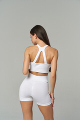 Athletic woman from behind in fitted white activewear, showcasing toned physique. She has long brown hair and is in a relaxed pose, emphasizing the back design of the outfit