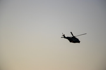 Silhouette of a helicopter flying at sunset with warm orange and red tones in the sky. Dramatic aviation scene on a colorful twilight background.

