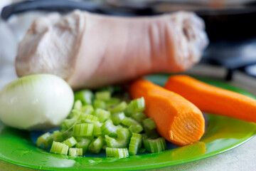 
A peeled pork shank is visible on a green plate, next to it is half an onion, chopped celery stalks, and two carrots.
