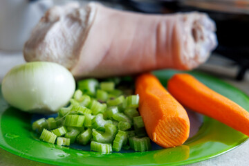 
A peeled pork shank is visible on a green plate, next to it is half an onion, chopped celery...