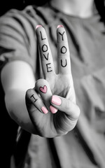 A black and white photo of a woman's hand shows a peace gesture with I Love You written on the fingers with a pink manicure and a heart.
