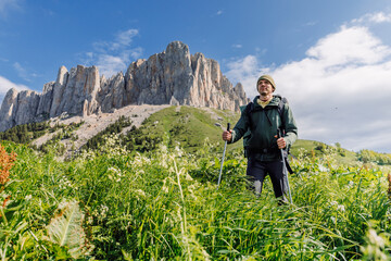 Male hiker with trekking poles stands in green meadow with rocky mountains
