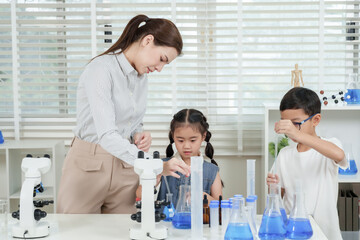 Caucasian female teacher teaching two asian elementary students performing science experiment together in classroom using microscope test tube beaker cylinder with blue liquid on white desk