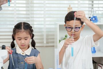 Asian elementary girl wearing denim top using pipette carefully in science class with blue chemical solutions on table in bright classroom full of lab equipment for learning experiment