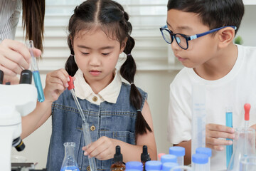 Asian girl using pipette to transfer blue liquid into beaker while boy watches carefully during...