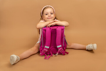 Little smiling girl sitting leaning on pink school backpack on beige background with space for...