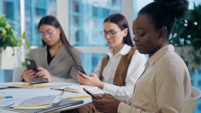 Modern multiethnic women females diversity team girls coworkers businesswomen business colleagues using mobile phone smartphone online app internet addiction scrolling social media indoors office