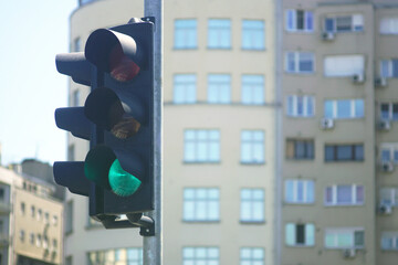 Urban traffic light showing green signal at city intersection with blurred modern residential buildings in background. Concept of safe driving, road regulation, navigation and transport infrastructure