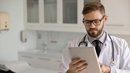 Serious young medical practitioner man in stylish eyewear and lab coat browsing histories on tablet, reading examination report. Doctor using digital gadget for communication