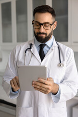 Positive handsome Hispanic doctor man in lab coat and eyewear thumbing through chats with patients...