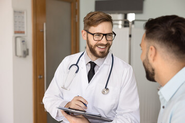 Candid portrait of male doctor talking to patient, writing medical records in survey form, holding...